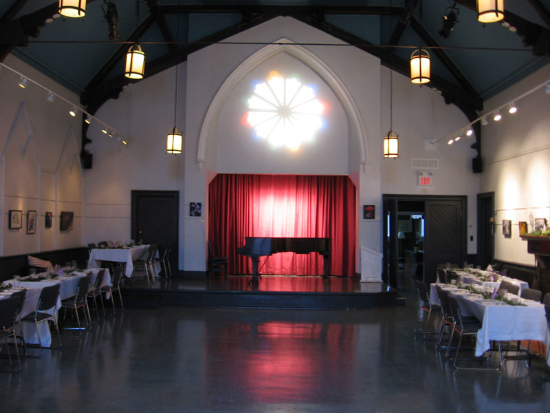 Front of main hall set up for a dinner event with chairs, tables, piano and club logo projected over the stage